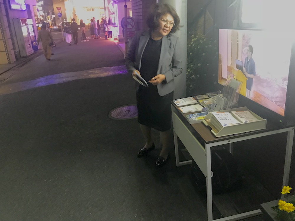 Emma Ballesteros, personal ministries director at Tokyo Central Seventh-day Adventist International Church, standing by a television showing a live-stream of an evangelistic meeting inside the church in Tokyo, Japan, on May 18, 2018. (Andrew McChesney / Adventist Mission)